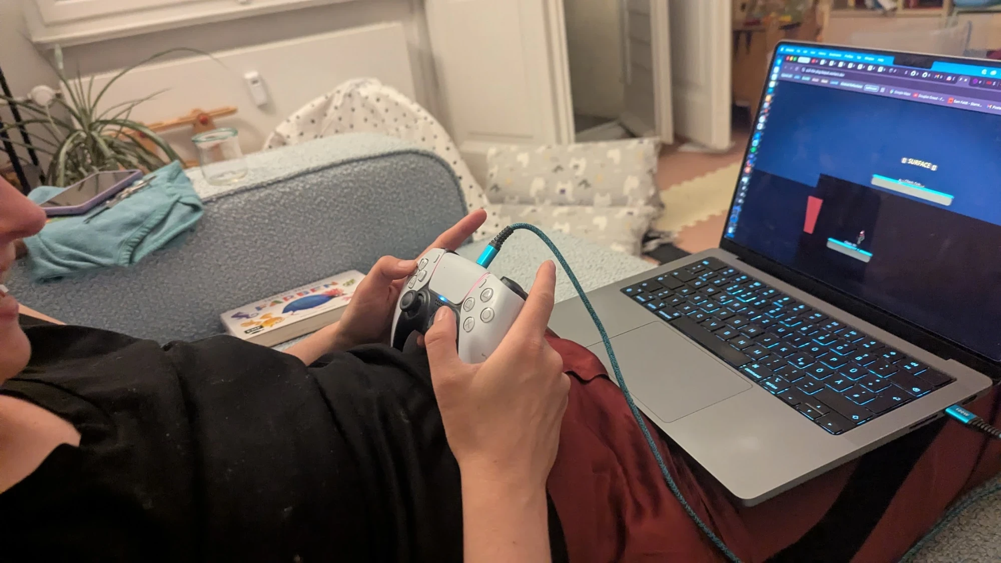 A first-person view of the couch: hands on a PlayStation 5 controller connected to a MacBook by a teal cable. On the screen, Mission 2 of Pull the Plug — the dark cave with the SURFACE marker label at the top, the player on a platform near rising water. A children's book sits on the couch beside them.